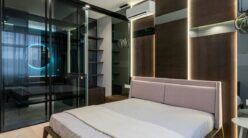 A modern bedroom with a minimalist design features a large bed with a light quilt, sleek wooden paneling, and ambient lighting. There are glass sliding doors, a mounted air conditioner, and a geometric shelving unit.