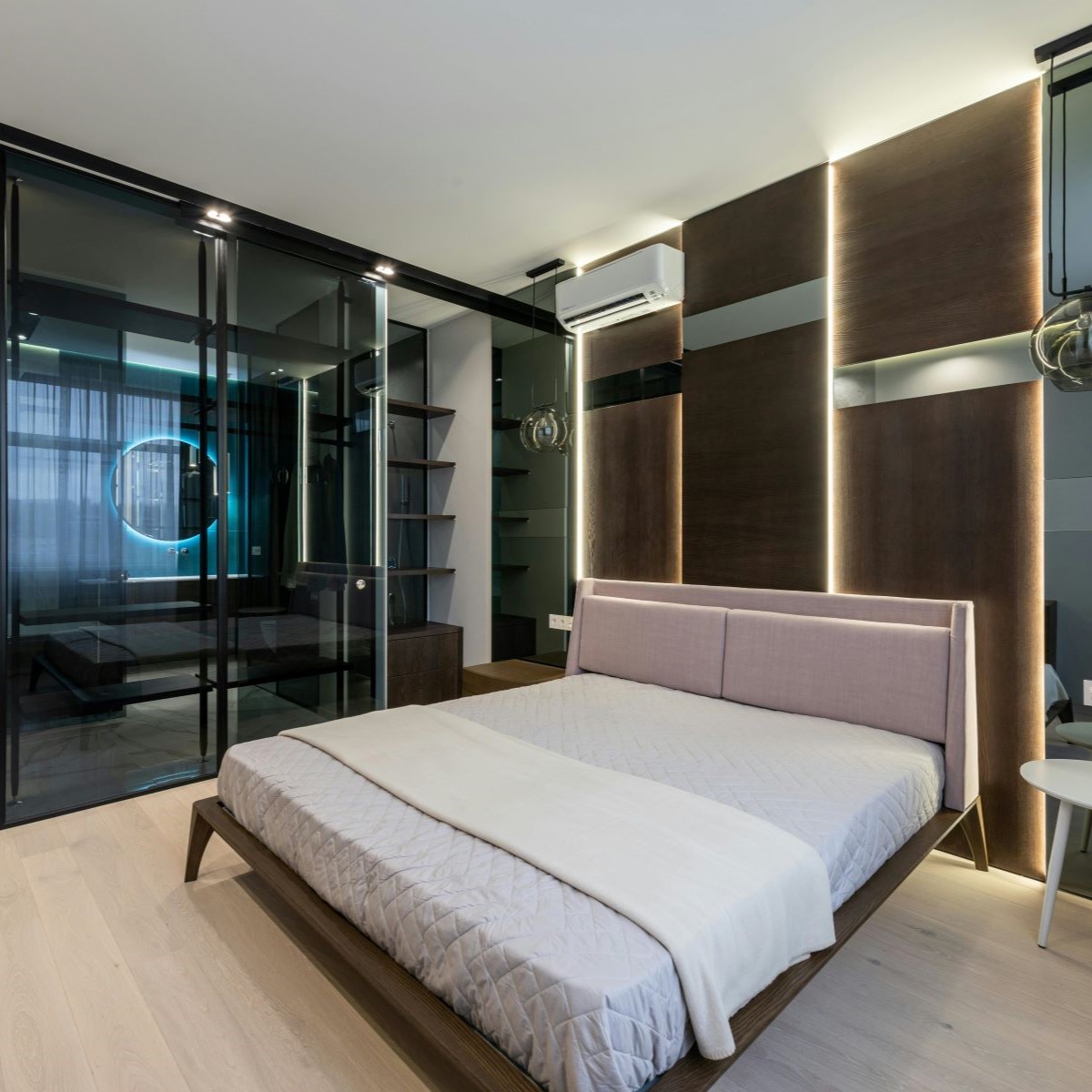 A modern bedroom with a minimalist design features a large bed with a light quilt, sleek wooden paneling, and ambient lighting. There are glass sliding doors, a mounted air conditioner, and a geometric shelving unit.