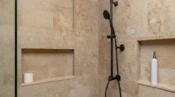 A modern shower with beige stone tile walls featuring a rainfall showerhead and a handheld showerhead. It includes built-in shelves holding a white soap dispenser and a roll of toilet paper. The setting appears clean and minimalist.