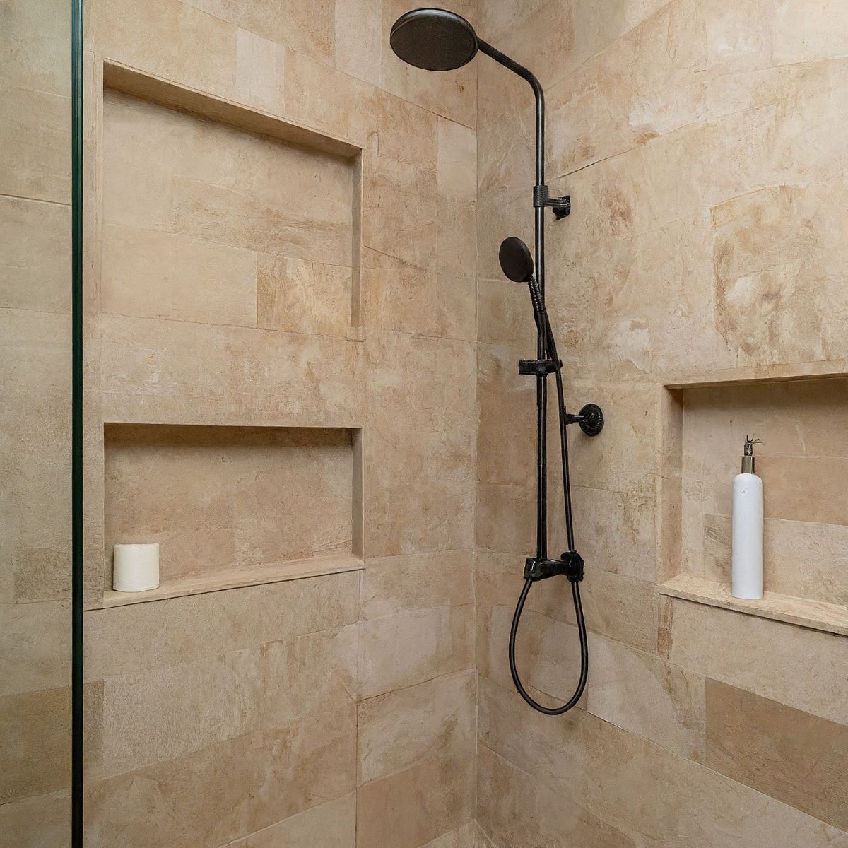 A modern shower with beige stone tile walls featuring a rainfall showerhead and a handheld showerhead. It includes built-in shelves holding a white soap dispenser and a roll of toilet paper. The setting appears clean and minimalist.