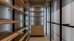 A modern, minimalist walk-in closet with wooden shelves lining both sides. The shelves are supported by sleek black metal frames. A small wooden cabinet is centered at the far end, and soft lighting illuminates the space.