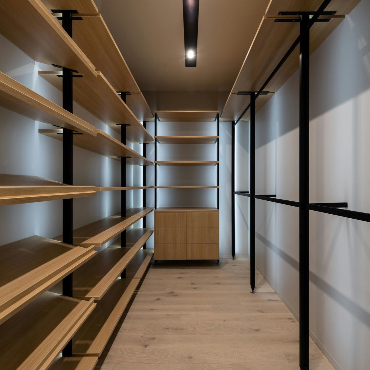 A modern, minimalist walk-in closet with wooden shelves lining both sides. The shelves are supported by sleek black metal frames. A small wooden cabinet is centered at the far end, and soft lighting illuminates the space.