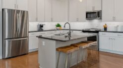 A modern kitchen featuring white cabinets, stainless steel appliances, and wooden flooring. A central island with a built-in sink has two wooden stools. Pendant lights hang above, and a plant decorates the counter.