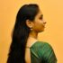 A woman with long, dark hair stands in profile against an orange background. She is wearing a green patterned top and decorative earrings.