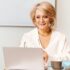 A woman with short blonde hair is sitting at a desk, smiling at a laptop. She is wearing a white blouse and a gold necklace. A cup and some notebooks are on the desk beside her.